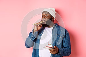 Happy Black hipster celebrating birthday, blowing party whistle, holding bday cake with candle, standing over pink