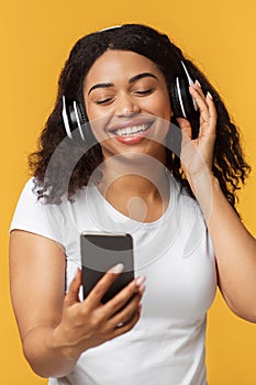 Happy black lady enjoying newest stereo headset and mobile music app, listening to music over yellow studio background