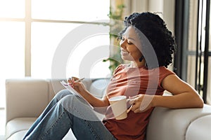 Happy black lady relaxing at home, drinking coffee and writing in notepad