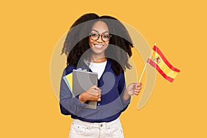 Happy black lady student with backpack and copybooks, holding Spanish flag, on yellow background