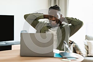 Happy black male freelancer looking at laptop computer and relaxing, holding hands behind head, sitting at desk at home