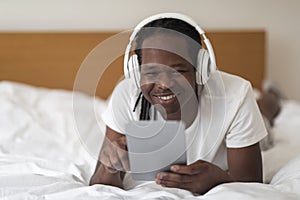 Happy Black Man With Digital Tablet And Wireless Headphones Relaxing In Bed