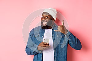 Happy Black man showing call me sign, making phone gesture and smiling, holding smartphone, standing in beanie and denim