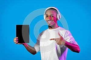 Happy black man showing digital tablet with mockup, using headphones