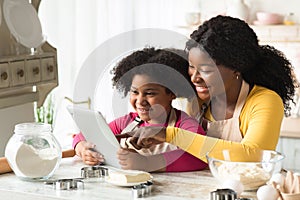 Happy Black Mom And Little Daughter Using Digital Table In Kitchen Together