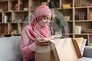 Happy black muslim female customer receiving long-awaited package, opening cardboard box, sitting on sofa at home