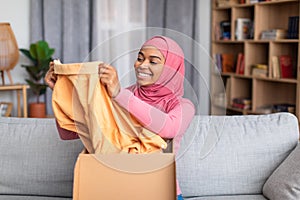 Happy black muslim woman opening carton box, checking her delivery, holding new clothes and smiling