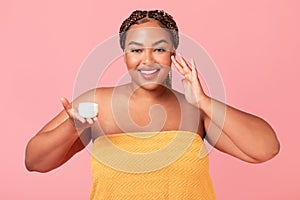 Happy black obese woman applying moisturising cream on face, standing wrapped in towel holding jar with cosmetic product