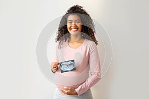 Happy black pregnant woman holding baby sonography photo and embracing belly