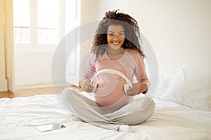 Happy black pregnant woman holding headphones near belly, playing melody to baby