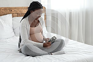 Happy black pregnant woman with ultrasound picture of baby sitting on bed