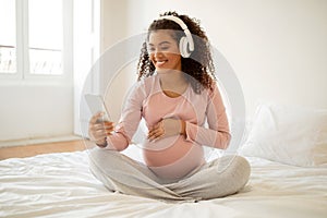 Happy black pregnant woman wearing headphones using smartphone at home