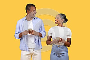 Happy Black spouses with cellphones, sharing smiles over a yellow background
