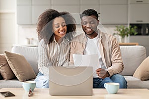 Happy black spouses paying bills via laptop holding papers indoor