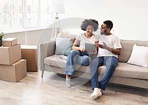 Happy Black Spouses Using Tablet Sitting Among Moving Boxes Indoor