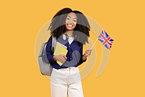 Happy black student with backpack, copybooks, holding Canada flag on yellow background, symbolizing language education