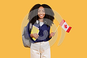 Happy black student with backpack, copybooks, holding Canada flag on yellow background, symbolizing language education