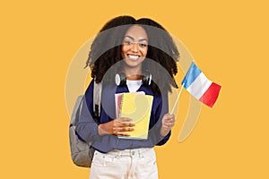 Happy black student with backpack and copybooks, holding flag of France, posing on yellow background