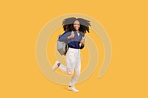 Happy black student lady with backpack and copybooks, joyfully jumping on yellow background