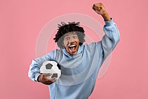 Happy black teenage guy with soccer ball rooting for his favorite football team on pink studio background