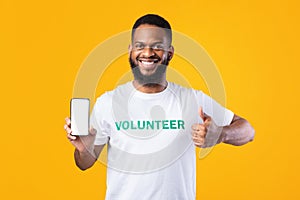 Happy Black Volunteer Showing Phone Screen Gesturing Thumbs-Up, Yellow Background