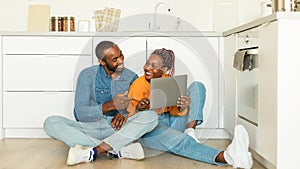 Happy black wife and husband using laptop, watch video or shopping online in kitchen interior, sitting together on floor