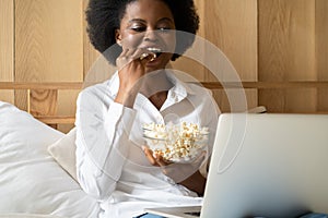 Happy black woman with afro hair style wear white shirt relaxing, eating popcorn while watching movie online on laptop, talking on