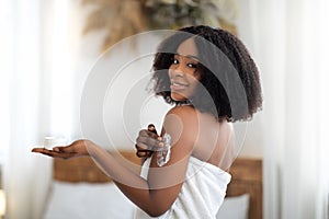 Happy black woman applying cream onto her body after shower, indoors