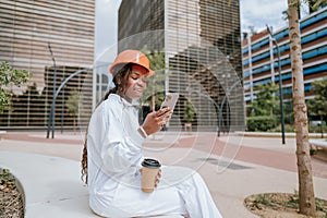 Happy black woman in helmet sitting with coffee cup and browsing smartphone