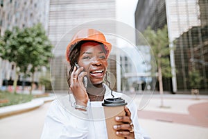 Happy black woman in helmet standing with coffee cup and speaking on smartphone