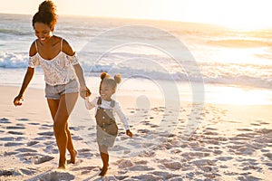 Happy black woman holding hands of daughter while walking on beach at sunset