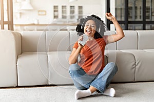 Happy black woman listening music and singing, sitting on floor at home