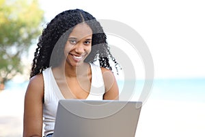 Happy black woman looks at you with a laptop on the beach