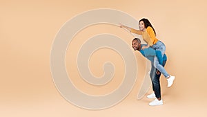 Happy black woman piggybacking her husband and pointing at free space, peach studio background, copy space, panorama