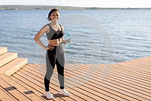 Happy black woman training next to lake, drinking water