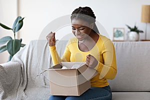 Happy black woman unboxing parcel at home, emotionally reacting to successful shopping photo