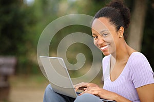 Happy black woman using laptop looking at you in a park