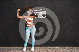 Happy black woman wearing face protective mask having fun and dancing while holding a retro radio - Focus on face