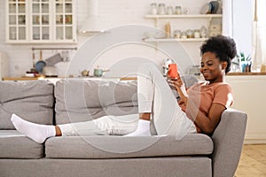 Happy Black young woman resting on cozy sofa at home, using smartphone, chatting in social networks.