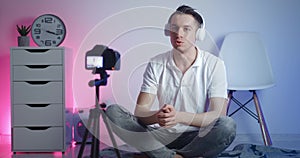 Happy blogger man in headphones filming new vlog video with professional camera while sitting on floor at home. Male