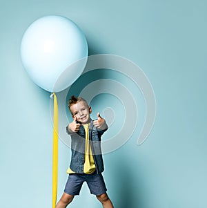 Happy blond boy in stylish casual clothing standing near blue air balloon and showing good thumb fingers sign over blue background