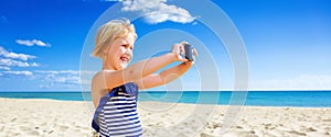Happy blond child on seacoast taking photo with digital camera