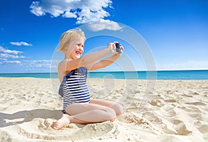 Happy blond child on seacoast taking photo with digital camera