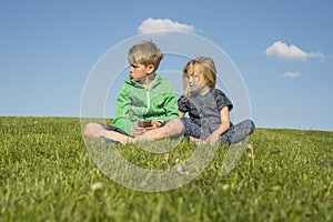 Happy blond children using smartphone (watching movie or playing game) sitting on the grass.