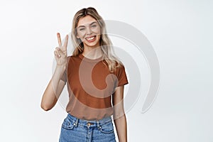 Happy blond girl shows peace, v-sign gesture and smiling, standing near empty copy space for advertisement, white studio