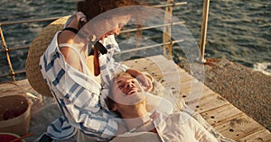 A happy blond guy with stubble in light clothes lies on the lap of his brown-haired girlfriend at a picnic on the pier