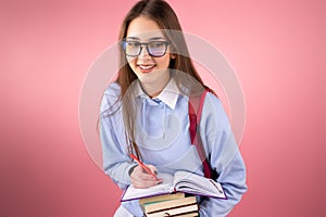 Happy blond high school girl in glasses smiling keeping stack of books and in a diary writing with a pen notes on pink