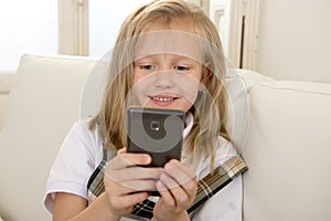 Happy blond little girl on home sofa using internet app on mobile phone