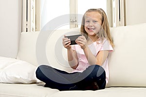 Happy blond little girl on home sofa using internet app on mobile phone