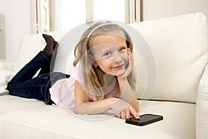 Happy blond little girl on home sofa using internet app on mobile phone